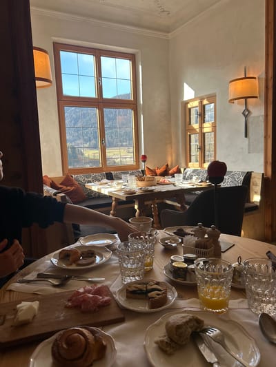 The image depicts a breakfast scene in Pflaurenz-Floronzo, Italy. A long wooden table is set for a meal, with a white linen cloth covering most of it. Dishes include plates of bread, pastries, and sliced meats. There are glasses of orange juice, water, and small jars of preserves. Seating consists of chairs on one side and a built-in cushioned bench on the other, adorned with orange pillows. A person, partially visible, is sitting at the table, their arm extended towards the dining setting. Large windows allow ample natural light into the room, framing a view of mountains in the distance. Lamps provide artificial light.