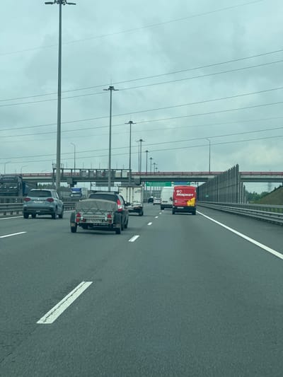 A wide-angle, eye-level shot captures a multi-lane highway under an overcast sky. The road surface is asphalt, marked with white lane dividers. Several vehicles are present: a blue Suzuki SUV with license plate E940XP 198 is in the left lane, followed by a dark car towing a trailer with license plate EE 4265 78. In the right lane, a red delivery van with the logo "04 Маркет" is visible, and further ahead, a white truck. Overhead, multiple lampposts line the highway, and a suspended bridge with a red railing crosses in the distance. Signs indicate exits for "МКАД Скандинавия" and "ПОРОШКИНО (POROSHKINO)", confirming the location in Poroshkino, Russia. A sound barrier made of grey panels runs along the right side of the highway.