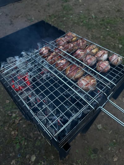This media file shows a close-up, overhead view of a metal grilling basket filled with food items, placed over a barbecue grill with glowing red coals and smoke. The food appears to be small, round pieces, possibly meatballs or sausages, each wrapped in strips of bacon. The bacon is visibly cooked, with some pieces appearing crisp and browned. The grilling basket itself is made of thin metal wires arranged in a grid pattern, and it has a handle. The barbecue grill is a dark, rectangular metal container filled with charcoal. The background is a blurry outdoor scene, with dirt, dried leaves, and some patches of green grass, suggesting a natural or garden setting. The lighting suggests it might be daytime, possibly late afternoon or early evening, as there are shadows. There is no visible text in the image. The scene evokes a sense of outdoor cooking and a casual gathering, with the focus on the food being prepared. Given the location context of Novaya Vozhoika, Russia, this scene likely depicts a common outdoor activity during warmer months.