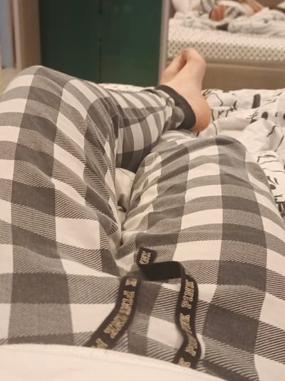 The image shows a close-up, low-angle view of a person's legs and feet resting on a bed. The person is wearing plaid pajama pants in shades of gray and white. A black drawstring with gold lettering that reads "PINK PINK PINK" is visible at the waist of the pants. The bed appears to be covered with a white sheet patterned with black designs. In the background, a blurred reflection in a mirror shows another bed with someone lying down. The environment suggests a domestic setting, likely a bedroom, and is not specific to Putilkovo, Russia.
