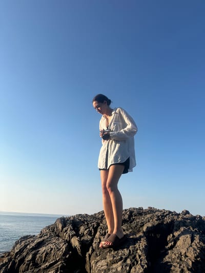 A young woman is captured in a vertical, low-angle shot, standing on a rocky outcrop with the vast expanse of a clear blue sky behind her. She is wearing a white, loose-fitting button-down shirt over dark shorts, and a pair of sandals. Her hair is pulled back in a bun, and she is looking downwards, holding a dark, cylindrical object in her hands, possibly a camera or a phone. Her posture suggests a moment of contemplation or focus.
The scene is set by the sea, with the calm, blue water visible in the lower right portion of the image. The rocks themselves are rough and textured, indicating a natural, coastal environment. The bright, clear sky suggests it is daytime, likely late afternoon or early morning due to the quality of light illuminating the woman's face and casting subtle shadows on the rocks. The overall mood is serene and tranquil, with a sense of connection to nature. There is no visible text in the image.