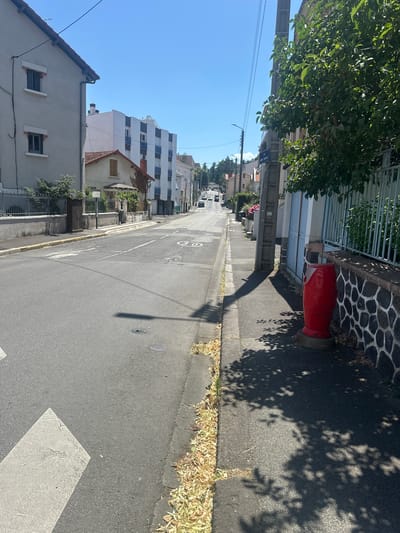 The image shows a street scene in Clermont-Ferrand, France. The street is paved and lined with buildings on the left and right. The buildings on the left are mostly residential, with some architectural variation. On the right, there is a tall utility pole, a fence, and a red cylindrical object. Further down the street, there are cars visible, and the road appears to be sloping upwards. The sky is clear and blue. A street sign reading "RUE ANDRÉ THURIOT" is visible on the right. There are some shadows from trees cast across the sidewalk and road.