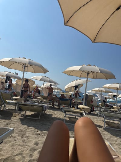 The image shows a sunny day at a crowded beach in Finale Ligure, Italy. The foreground is dominated by the perspective of someone lying on a beach chair, with their legs in the frame. The middle ground is filled with rows of beige and white beach umbrellas and lounge chairs. Several people are visible, some sitting under umbrellas, others walking or standing. In the background, the calm sea and clear blue sky are visible. A flag, possibly the Italian flag, can be seen on a pole among the umbrellas. The beach is sandy, with visible footprints and shadows cast by the umbrellas.