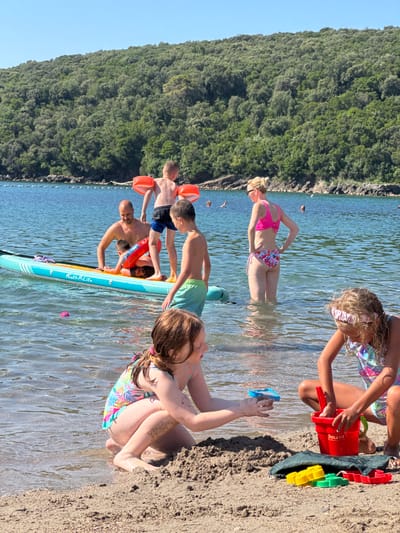 The image depicts a group of people at a beach in Prijevor, Montenegro. The scene is set on a sunny day with clear blue water and a lush, green forested hillside in the background. Several individuals, including adults and children, are interacting in and around the shallow water.

In the foreground, a child with blonde hair is partially submerged in the water, reaching for a blue toy. Next to them, a woman in a pink bikini stands in the water, her hand on her hip. A group of boys are gathered on a stand-up paddleboard, some holding bright orange flotation devices. One boy is reaching towards a red bucket filled with sand toys. Further back, another child is sitting on the sandy shore, also playing with sand and toys.

The overall mood appears to be relaxed and joyful, indicative of a family outing or a day at the beach. The water is calm and reflects the sunlight, creating a shimmering effect. Visible text on the paddleboard reads "Feath-R-Lite". The presence of sand toys suggests children are engaged in typical beach activities. The time of day is likely midday, given the bright sunlight and shadows.