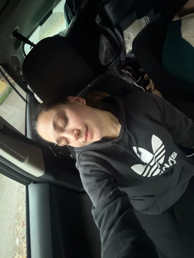 Within the interior of a vehicle in Lonate Pozzolo, Italy, a young female is observed occupying a rear passenger seat, appearing to be asleep. She is positioned with her head tilted, eyes closed, and exhibits a relaxed facial expression. The individual is dressed in a dark grey or black hooded sweatshirt featuring a white Adidas Trefoil logo on the chest, paired with dark-colored pants.

Behind the front passenger seat, a black object resembling a long case with a protruding handle is visible, extending vertically. Adjacent to this, various dark-colored items, possibly clothing or bags, are present in the rear compartment. A partial view of another individual's clothing, specifically teal and white fabric, is discernible in the upper right quadrant of the image, suggesting the presence of at least one other occupant. Through the left-side vehicle window, a blurred exterior environment is apparent, showing a wet, textured surface consistent with a paved road or lot and some scattered light-colored debris. The lighting indicates a daytime setting.