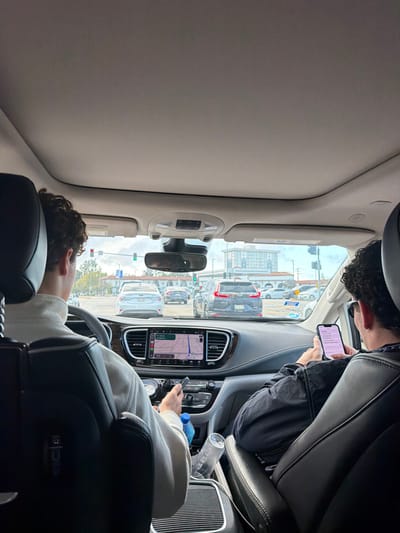 Inside a car, the driver is resting their head on the seat while looking at a phone in their hand. A passenger is in the seat above them, also looking at a phone. The car seems to be stopped in traffic, as cars are visible outside the windshield. The car's navigation system is on, displaying a map.