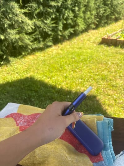 In Viden, Bulgaria, on a sunny day, a person's hand, adorned with yellow-painted fingernails, holds a dark blue tobacco heating device. The device is partially inserted into a tobacco stick. The hand rests on a colorful striped towel, predominantly yellow, red, and white, with a blue towel folded underneath. The background features lush green grass and the dappled shade of trees, suggesting an outdoor setting, possibly a garden or park.
