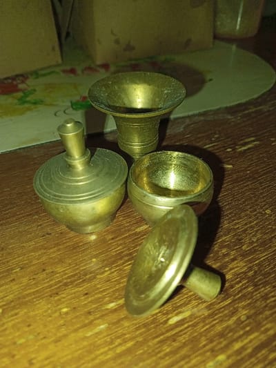 The scene is set indoors in Longmiang, Laos, on a wooden surface displaying distinct grain patterns and signs of wear. Three small, aged metallic objects, appearing to be brass or bronze due to their color and tarnish, are arranged closely together.

The object on the left is a small, lidded vessel with a rounded body and a pointed finial on its slightly-askew lid. To its upper right, a trumpet-shaped vessel stands upright, featuring a flared rim and a narrower base. Directly below and to the right of the trumpet-shaped vessel, an open, small, spherical bowl rests. Adjacent to this open bowl, on the lower right, lies a separate disc-shaped lid with a cylindrical stem or handle, positioned on its side. All metallic items exhibit visible tarnish and surface irregularities.

The background is blurred, revealing a light-colored surface with a faint floral or patterned design behind the objects, and a rectangular, light-brown object further back on the left. Illumination appears to be from an artificial light source, casting subtle shadows. There are no people or discernible actions.