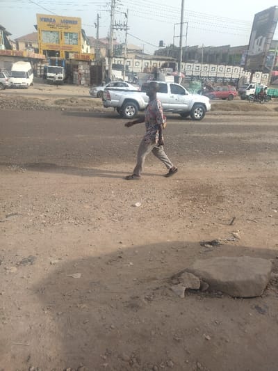 The image depicts a man walking across a gravel road in Way, Nigeria, located at latitude 9.029 and longitude 7.584. The man is wearing a light-colored shirt with a floral pattern and light-colored pants. He is walking to the right, away from the camera. He is carrying a dark-colored bag on his right shoulder. In the background, there are multiple vehicles parked and driving on the street. The road is partially covered in gravel and dirt. The image appears to have been taken on a sunny day. There are buildings and shops on the other side of the road.