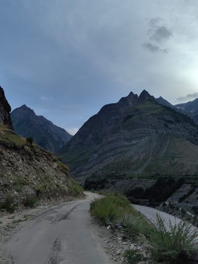 The image depicts a mountainous landscape with a dirt road winding through it. The road is in the foreground, appearing rough and unpaved, with patches of grass growing along its edges and in the middle. On the left side of the road, there is a steep, rocky embankment covered in sparse vegetation.

In the mid-ground, the road curves towards the right, leading into a valley. A river with murky water flows through the valley floor. Steep, green, and rocky mountains rise on either side of the valley. The mountains are characterized by layered rock formations and vegetation. Several peaks are visible in the background, some with jagged summits.

The sky is overcast with scattered clouds, creating a diffused light over the scene. The overall impression is one of a remote and rugged natural environment. The location is identified as Gushal, India, suggesting this is a scene from that region. There are no people or animals visible in the image.