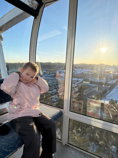 A young child is depicted inside a spacious, enclosed Ferris wheel gondola in the city of Mistolovo, Russia, during what appears to be a clear winter sunset. The child, approximately 5-8 years old and wearing a light pink winter jacket and dark grey winter pants, is seated on a blue patterned bench seat. She is facing the camera with her hands positioned under her chin, exhibiting a slight smile. The gondola features large, clear glass windows framed in white, through which a snow-covered urban-suburban landscape is visible. This exterior view includes buildings, roads, numerous bare trees, and a large parking lot densely populated with vehicles. The low-angle sun is prominent in the upper right quadrant of the image, causing a bright glare on the window glass and illuminating the scene with warm light. Contrails are discernible in the clear blue sky. Several worn or damaged informational stickers are affixed to the lower section of the windowpane. The gondola floor is textured grey metal.