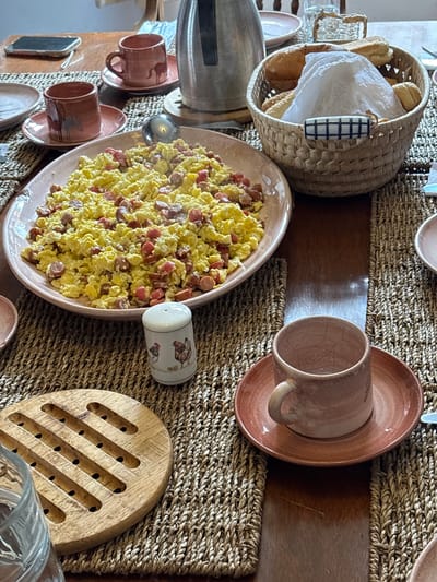 The image depicts a breakfast setting on a wooden table, likely in Aucallama, Peru. A large platter of scrambled eggs with sliced hot dogs is centrally placed. To the left, two small terracotta cups and saucers with horse figures are visible, one holding a hot beverage. A woven basket filled with bread rolls and covered with white paper sits to the right of the egg platter. In the foreground, a rustic wooden trivet and a glass of water are present. A salt shaker with chicken illustrations is also on the table. The overall ambiance suggests a casual, home-style meal.