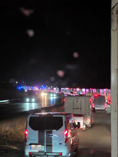 The image depicts a nighttime traffic scene on a highway, likely near Uettingen, Germany. A line of vehicles, including vans and trailers, are stationary or moving slowly, indicating a traffic jam. The road surface appears wet, reflecting the headlights of the vehicles and the lights from a distant structure that may be a toll booth or service area. Several vehicles are illuminated with flashing blue and red lights, suggesting the presence of emergency services. The sky is dark, with a few blurry lights visible, possibly from streetlights or distant buildings. The foreground shows the rear of a white van. The overall atmosphere is one of disruption and potential incident due to the emergency lights and traffic congestion.