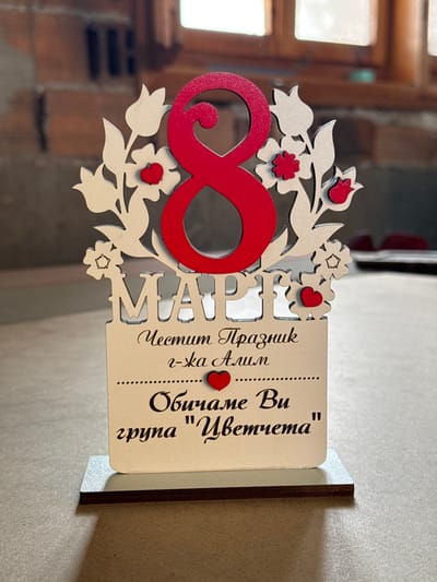 The image displays a laser-cut wooden decorative plaque positioned on a light-colored surface, likely a tabletop or workbench. The plaque is a gift for International Women's Day, March 8th, indicated by the prominent red numeral "8" at the top center, surrounded by white floral and foliage designs with small red flower accents and hearts. Below the number, the word "МАРТ" (MARCH) is cut out in white. The lower section of the plaque, also white, bears text in black script: "Честит Празник г-жа Алим" (Happy Holiday Mrs. Alim), followed by a dotted line, a red heart, another dotted line, and then "Обичаме Ви група "Цветчета"" (We Love You group "Little Flowers"). The plaque stands on a darker, thin rectangular wooden base.
The background is out of focus, revealing an indoor setting. To the left, a reddish-brown brick wall or structure is partially visible. To the right, a window with a visible wooden frame allows bright natural light into the scene. The object is located in the city of Breznitsa, Bulgaria.