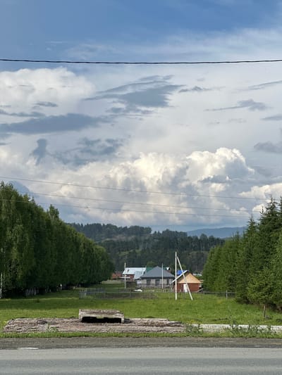 The image depicts a rural scene in Inzer, Russia, under a dramatic sky. In the foreground, a grey asphalt road runs horizontally, with a patch of rough ground and concrete debris to its right. Behind the road is a grassy field, interrupted by a large, weathered concrete slab with visible rebar. A wooden fence runs along the edge of the field, separating it from a row of lush green birch trees on the left and dense evergreen trees on the right.

Further into the scene, several houses with distinct roofs are visible in the mid-ground. One house has a white roof and what appears to be light-colored walls, while another nearby structure is smaller and painted red with a light-colored triangular roof. A wooden utility pole stands in front of these houses. The background is dominated by a rolling, tree-covered hillside under a sky filled with a mix of bright white cumulus clouds and darker, more diffused grey clouds. The overall impression is one of a quiet, rustic village setting on a day with changeable weather. There are no people visible in the image.