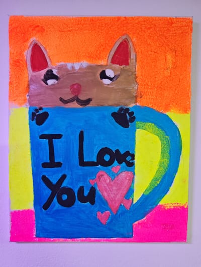 A colorful painting depicts a cartoon cat peeking out of a blue mug. The cat has brown fur, red ears, and black eyes with white pupils. Its nose is pink, and it has black whiskers. The mug has black paw prints on it, and the words "I Love You" are written in black lettering inside the mug. Three pink hearts are also depicted on the mug. The background of the painting is a vibrant orange, yellow, and pink. This painting is located in Memmingen, Germany.