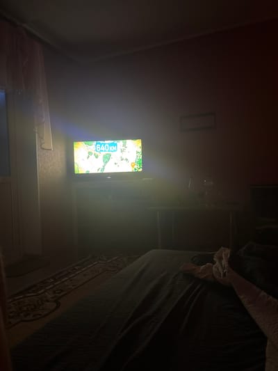 A dimly lit bedroom in Sturzeni, Moldova, contains a large flat-screen television displaying a map with the text "640KM" prominently featured in a blue box. The television is placed on a stand in front of a wall. To the right of the television, a desk or table is visible, holding various objects, including a bottle and a microphone. A bed with dark bedding occupies the foreground of the image, with a patterned rug on the floor to the left. A window with white curtains is present on the far left side of the room. The overall atmosphere is dark, with the television screen being the primary light source.