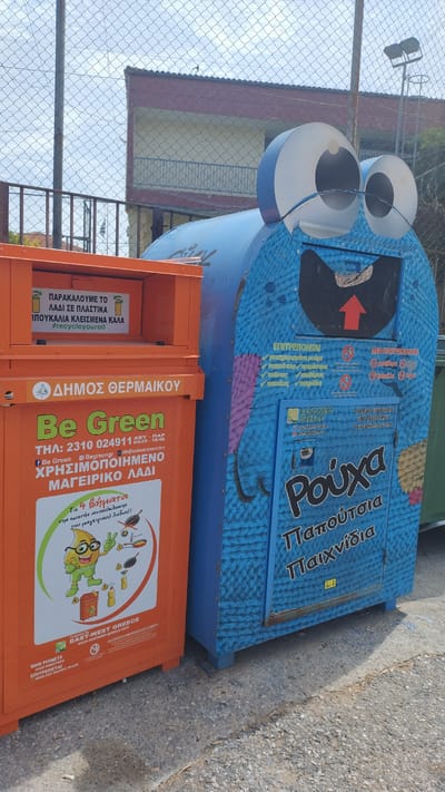 The image shows two recycling bins in Νέα Μηχανιώνα, Greece. The bin on the left is orange and has text in Greek indicating it is for the Municipality of Thermaikos and is for cooking oil. It also has a cartoon character promoting recycling. The bin on the right is blue and shaped like a monster with large googly eyes. It has text in Greek indicating it is for clothes, shoes, and toys. Both bins appear to be located outdoors, near a fence and a building.