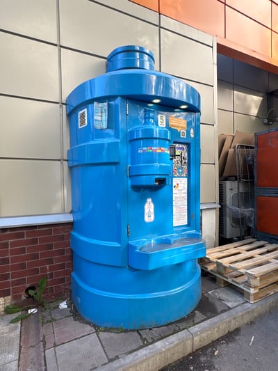 A bright blue water dispensing machine, resembling a large stylized water bottle, stands on a paved sidewalk next to a brick wall and a building facade. The machine has a cylindrical body with horizontal bands and a domed top. It features illuminated buttons, a coin slot, and a dispensing area with a platform for placing a bottle. Various labels and instructions are visible on its front panel, including a phone number and price information in Russian.

To the right of the water dispenser, a stack of cardboard boxes is visible, along with metal shelving and what appears to be an air conditioning unit or heat exchanger. Further to the right, a large red industrial container is partially visible. The ground is a mix of paved stones and concrete. A small patch of green weeds is growing near the base of the brick wall. The overall scene suggests an outdoor service area, possibly near a commercial building. The location is identified as Bykovo, Russia.