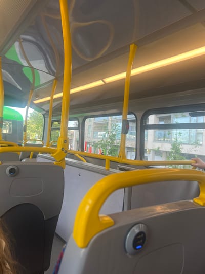 The image captures the interior of a public bus, likely in Bray, Ireland, given the context. The scene is dominated by yellow grab rails, a common feature in public transportation, which run along the ceiling and sides of the bus. The bus appears to be in motion, as suggested by the blurred reflections in the windows and the dynamic perspective of the interior.
Through the windows, buildings and trees are visible, indicating an urban or suburban setting. The time of day appears to be daytime, with natural light filtering in, illuminating the interior with a warm, yellowish glow from the overhead fluorescent lights. The weather is not explicitly determinable from the image, but the bright interior lighting suggests it's not heavily overcast.
The focus of the image is on the bus's interior elements, particularly the yellow handrails and the grey seating or paneling. A subtle detail is a USB charging port with a blue indicator light, visible on a grey panel near the top right of the frame, suggesting modern amenities on the bus. No people are clearly identifiable as subjects, though a partial view of a hand holding a grab rail is present at the top right. The overall impression is that of a typical commute or journey on a bus. No discernible text is visible in the image.