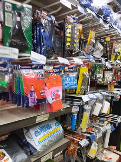 The image presents a busy aisle of a retail store, filled with various packaged products displayed on white shelves and hanging racks. The setting suggests a general merchandise, hardware, or automotive accessory shop. No people are visible, but a graphic of a person wearing a rain poncho appears on one of the product packages.

The shelves are densely stocked with a diverse range of items. Visible products include bicycle accessories like seats and handlebar grips (yellow and black striped), blue and orange gloves, rain ponchos, and various tools such as wrenches. There are also flashlights (blue and yellow) and other small packaged items, possibly household or vehicle maintenance accessories. Most items are individually packaged in clear plastic, often hanging from hooks.

Notable details include the repetitive "PITSTOP" brand visible on many packages. A prominent silver package on a lower shelf reads "Elmondo jas hojen 2 in 1 HELIOS," with "jas hojen" translating to "raincoat" in Indonesian. This, along with price tags such as "13.000," "5.000," and "125.000," suggests a currency style common in Indonesia (Rupiah) and the presence of Indonesian language, which contrasts with the provided location context of Aglipay, Philippines. Another visible text is "Ho" on a glove and "HEAD" and "push" on other packages. The overall scene depicts a functional retail space designed for product display and sales.