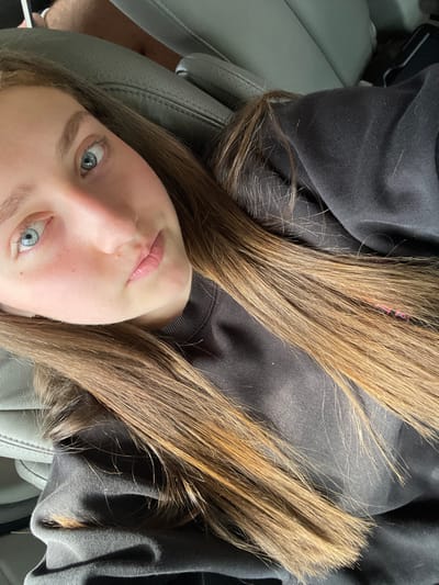 A young woman with light blue eyes and fair skin reclines on a gray leather seat. Her long, straight, light brown hair fans out around her head and across her dark sweatshirt. The seat appears to be in a car, as a section of another seat is visible in the background. The image is taken from a slightly high angle, looking down at her face.