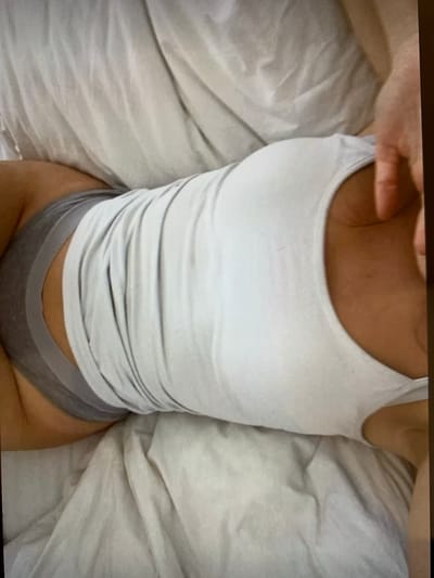 The image shows a person lying on a bed with white sheets. The person is wearing a white tank top and grey underwear. A hand is visible on the right side of the frame, resting on the person's shoulder. The scene is indoors. There is no information about the location in the city of Konstantinovka, Kyrgyzstan.