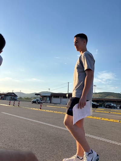 A young man stands outdoors on a paved surface, possibly a parking lot, holding a document. He is wearing a gray t-shirt, black shorts, and white sneakers. In the background are buildings, vehicles, and a mountain range under a clear sky. The location is Dimitrovgrad Municipality, Serbia.