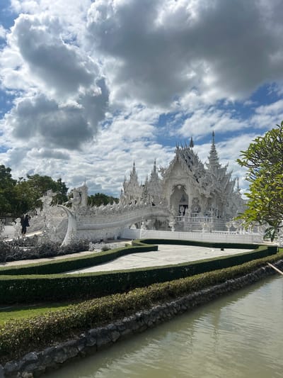 This detailed image depicts Wat Rong Khun, also known as the White Temple, located in Ban Rong Khun, Chiang Rai Province, Thailand, under a partly cloudy sky. The central subject is the temple's highly ornate main building, an entirely white structure featuring intricate traditional Thai architectural elements such as multi-tiered roofs, elaborate gables, and spires. Its surfaces are rich with sculptural details and reflective segments. A distinctive white bridge, flanked by complex railings and an array of sculptural elements including outstretched hands emerging from a lower area, extends towards the temple's primary entrance. In the foreground and to the right, a body of tranquil water reflects the sky, bordered by a low stone wall and vibrant green landscaping. Manicured dark green hedges delineate pathways across the temple grounds. A single individual dressed in dark clothing stands on the left side of the bridge structure, oriented towards the temple. Additional faint figures are discernible near the main temple entrance, indicating visitor presence. Dense green foliage from trees is visible on the far left, while a lighter green leafy branch partially frames the upper right corner of the scene.