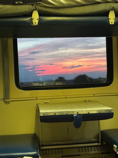 The image shows a window view from a train carriage, looking out at a vibrant sunset. The sky is filled with dramatic clouds painted in hues of pink, orange, and purple, contrasting with the darker blue of the upper sky. Silhouetted trees and a distant landscape are visible beneath the colorful sky. Inside the train, the view is framed by the yellow interior walls and the dark blue upholstery of the seats. A folded-down tray table with a blue fabric skirt is visible below the window. The image does not provide any information about the city of Halkidon, Russia, or any specific details about people or actions.