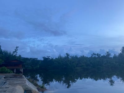 The image depicts a serene twilight scene overlooking a body of water with a dense forest lining the opposite bank. In the foreground, a concrete structure with stairs descends towards the water. To the left, a wooden stilt house with an orange roof and striped railings is partially visible, nestled among foliage. The water's surface acts as a mirror, reflecting the surrounding trees and the cloudy sky. The sky itself is a gradient of deep blues and grays, with scattered clouds suggesting the lingering light of dusk or the approaching dawn. The environment appears to be natural and tranquil, likely situated in a rural or semi-rural area. Given the context provided, this scene is located in Aneuk Paya, Indonesia, characterized by its lush vegetation and proximity to water. There are no people visible in the image.
