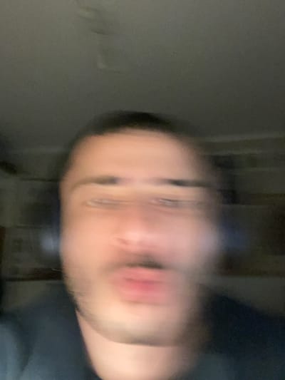 The image features a close-up, severely motion-blurred shot of a person's face. The individual appears to be an adult, possibly male, with visible stubble or a short beard around the chin and jawline. Their face is angled, with the head tilted, and the eyes are somewhat discernible despite the blur. The mouth is slightly open, revealing reddish lips. Due to the significant blur, detailed facial features and expressions are indistinct. The background is dark, out of focus, and featureless, suggesting an indoor setting with low ambient light. Some faint vertical lines or shadows are present in the background, but they are too blurry to identify. There are no distinct objects or discernible text in the image. The low light conditions suggest the photo might have been taken in the evening or at night, or in a dimly lit room. The scene is set in Ganei Tikva, Israel.
