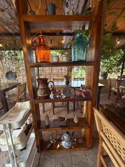 The image displays a detailed view of an indoor or semi-outdoor dining establishment in Sanctorum, Mexico. The primary subject is a tall, five-tiered wooden shelving unit positioned centrally in the foreground.

**Objects on the Shelf (from top to bottom):**
*   **Top Shelf:** A dark gray cylindrical mug is on the left. A small, light-colored, possibly ceramic, animal figurine is on the right.
*   **Second Shelf:** An orange, textured glass bottle with a handle is on the left. Two small, cylindrical, cream-colored jars with labels are in the center. A teal-blue, textured glass bottle with a handle is on the right.
*   **Third Shelf:** A brown ceramic pitcher with red, green, and white geometric patterns is on the left. A small, reddish-brown terracotta figurine resembling an animal (possibly a dog or boar) is next to it. A light-colored, patterned, decorative gourd or pumpkin-like object is in the center. Another reddish-brown terracotta figurine, elongated and textured, resembling an armadillo or similar creature, is on the right.
*   **Fourth Shelf:** Two elongated, light-colored, textured objects resembling corn cobs are on the left and right sides. A small, dark, rectangular object rests between them.
*   **Bottom Shelf:** A piece of gnarled driftwood or root is on the left. A blue and white ceramic teapot with a detailed floral or geometric pattern (reminiscent of Talavera pottery) is in the center. A small, flat, light-colored stone is on the right.

**Surrounding Environment:**
*   **Flooring:** The floor consists of irregularly shaped, light gray stone tiles.
*   **Background Seating:** Multiple wooden dining tables and chairs with woven or slatted backs are visible in the mid-ground and background, suggesting a seating area. One table has a small black vase with colorful dried flowers.
*   **Walls/Screens:** A natural bamboo or reed screen wall serves as a partition or backdrop on the left and center, allowing glimpses of lush green foliage beyond.
*   **Ceiling/Roof:** An overhead wooden structure, possibly a pergola or roof, is partially covered with interwoven straw/thatch material and green leafy vines. String lights with exposed bulbs are suspended across this structure.
*   **Foreground (Left):** A white, two-tiered metal rolling utility cart is positioned to the left of the wooden shelf. Its shelves hold stacks of neatly folded white cloth napkins or towels.

**People, Actions, Interactions:**
*   No people are visible within the frame. There are no ongoing actions or interactions depicted.

The scene suggests an unoccupied, inviting, and naturally lit restaurant or cafe space, decorated with traditional and rustic elements.