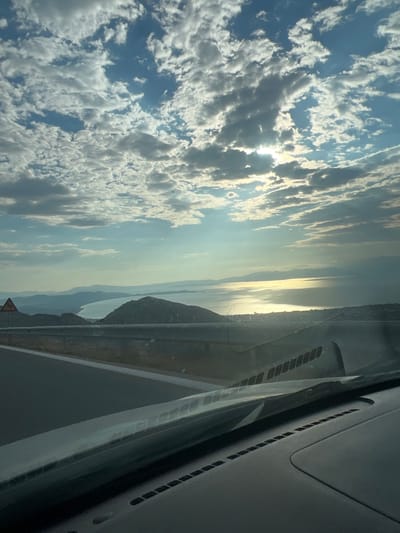 The image is a vertical, eye-level, wide shot captured from inside a vehicle looking out at a scenic coastal view under a cloudy sky. In the foreground, the dark dashboard of the vehicle is visible, with the windshield partially obscuring the view. A road curves to the left, with a guardrail separating it from a steep drop-off.
Beyond the road, a dark, hilly landscape stretches out, leading to a body of water that reflects the bright, sunlit clouds. The sun is partially obscured by clouds, creating a shimmering effect on the water. In the distance, layers of hazy mountains recede into the horizon. A small, dark object, possibly a bird or drone, is visible in the sky above the distant mountains. A yellow triangular warning sign with a black zigzag symbol is present on the left side of the road.
The scene appears to be located in or near Nea Makri, a municipal unit within the Marathon municipality of Attica, Greece, known for its coastal beauty and hilly terrain. The overall atmosphere is serene and expansive, highlighting the natural landscape under a dynamic sky. There are no people visible in the image.