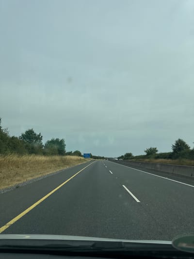 The image shows a view from inside a vehicle traveling on a highway in Ireland. The highway is paved with asphalt and has a yellow line separating the lanes. The sky is overcast and the landscape consists of dry grass and trees on either side of the road. There is a blue highway sign visible in the distance to the right. There are no people or other vehicles immediately visible in the foreground. The location is not specifically identifiable as Rivergrove, Ireland from the image alone.