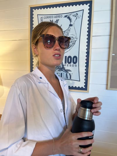 An indoor portrait features a young adult, likely female, holding a black thermos or water bottle with both hands. She wears large, tortoiseshell-patterned sunglasses, obscuring her eyes. Her mouth is slightly open, revealing teeth. She is dressed in a white collared shirt, partially unbuttoned to show a grey undergarment and a thin necklace. A small embroidered star design is visible on her shirt collar. She wears a ring on her left ring finger and a delicate bracelet on her right wrist.

In the background, a large framed piece of art, designed to look like a vintage postage stamp, is prominently displayed against a white, horizontally paneled wall. The stamp features an illustration of a bird, possibly a Rhea, and text elements such as "CORREO AEREO," "100 PESOS," and "UGUAY" (likely part of "URUGUAY"), strongly hinting at the location of Uruguay, specifically Faro de José Ignacio as per the context. The thermos she holds has a silver and black cap and a partially visible brand name "MUT" or "MUTTU." The lighting suggests it is daytime.