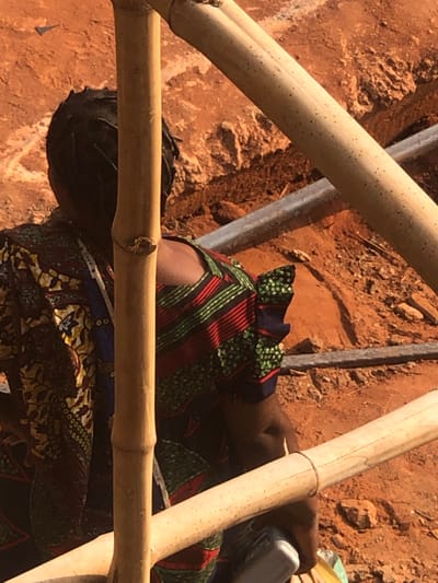 The scene depicts a solitary individual observed from behind, partially obscured by bamboo scaffolding, at a construction or excavated site in Agukwu Nri, Nigeria. The individual, dressed in a multi-patterned garment featuring horizontal green, red, and black stripes on the upper portion and a darker fabric with yellow, blue, and white patterns on the back, is positioned low to the ground, possibly kneeling or stooping. Their dark hair is visible at the back of the head. A light-colored, possibly rectangular object is held in their right hand. The environment consists of exposed reddish-brown earth, indicative of disturbed ground with visible depressions and trenches. Two silvery-grey linear objects, resembling metal rods or pipes, lie horizontally on the ground in the background. Bright sunlight illuminates the scene, casting minimal shadows.