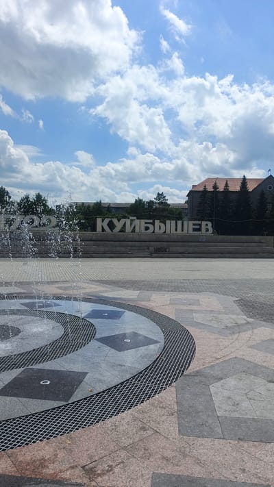 The image shows a public square with a fountain in the foreground and large white letters spelling out "КУЙБЫШЕВ" in the background. The fountain is spraying water, creating a dynamic effect. The square is paved with stone tiles of various colors and patterns. Behind the fountain, there is a tiered structure where the large letters are placed.

The letters are positioned in front of a row of trees and buildings. One building visible behind the letters has a red tiled roof and is made of stone. The sky above is blue with scattered white clouds, suggesting it is daytime and the weather is fair. The scene appears to be a calm and pleasant day in an urban environment.

The visible text in the image is "КУЙБЫШЕВ", which is the name of the city (Kuybyshev, Russia). The letters are large and three-dimensional, serving as a prominent landmark or monument.

No people are visible in the image, and the focus is on the architectural elements and the natural setting. The overall impression is one of a civic space, likely a central square or park, designed for public enjoyment.