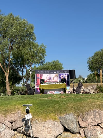 The image captures an outdoor event setup against a backdrop of natural elements, possibly in or near Pueblo Nuevo de Jasso, Mexico. A promotional display, likely for a tourism or travel-related company ("VIDANTAWORLD Vacation Planner"), features images of attractions such as a Ferris wheel and performers. An audio speaker is positioned near the display, suggesting a presentation or performance is planned. A camera on a tripod is set up, with a sign reading "NO BLOQUEAR CAMARA" (Do not block camera), indicating the event is being recorded or livestreamed. The scene is set on a grassy area, bordered by rocks and foliage, with a clear blue sky overhead. A tree stands prominently nearby, adding to the outdoor atmosphere. The presence of equipment bags suggests recent setup or ongoing activity.