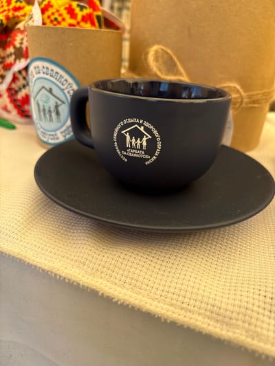 A close-up shot focuses on a dark blue coffee cup and saucer placed on a textured beige cloth. The cup bears a white circular logo with text and a silhouette of a family under a house, accompanied by a steaming cup. The text in the logo reads "СЕМЕЙНОГО ОТДЫХА И ЗДОРОВОГО ОБРАЗА ЖИЗНИ" and "«ГАРБАТА ПА-СВАЯКОЎСКI»". In the background, a cardboard container with a similar blue logo is visible, filled with colorful fabric. Another larger, light brown cardboard container with twine wrapped around it is also in the background. The scene is lit by warm light, casting a shadow of the cup and saucer onto the cloth. The image is taken from a slightly elevated angle. The location is identified as Вяйнянскі сельскі Савет, Belarus.