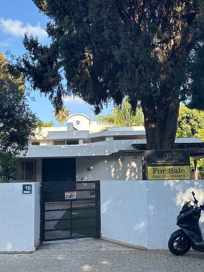 This image, taken during the daytime under a clear blue sky with scattered clouds, displays the exterior of a residential property. A prominent "For Sale" sign is visible, reading "R Real Estate Consultant Entrepreneurship & Investments For Sale Eyal: 054-5245972". The sign is affixed to a pole near a large, ancient-looking tree with dense green foliage that dominates the left side of the frame.

The house itself is predominantly white, featuring dark-framed windows and a sturdy black metal gate at the entrance. On the white wall beside the gate, a number plate clearly shows "16 קורסיא" (16 Kursia), identifying the house number and street name. A smaller, red and white sign on the gate warns "זהירות כלב!" (Caution Dog! in Hebrew).

A black scooter is parked on the light-colored, patterned paved area in front of the house, against a white wall. The scene is well-lit, with distinct shadows cast on the house and ground, indicating direct sunlight. Various green trees and bushes surround the property, adding to a lush environment, including what appears to be a palm tree in the background. The visible Hebrew text and phone number with an Israeli prefix suggest the location is in Israel, specifically the street Kursia.