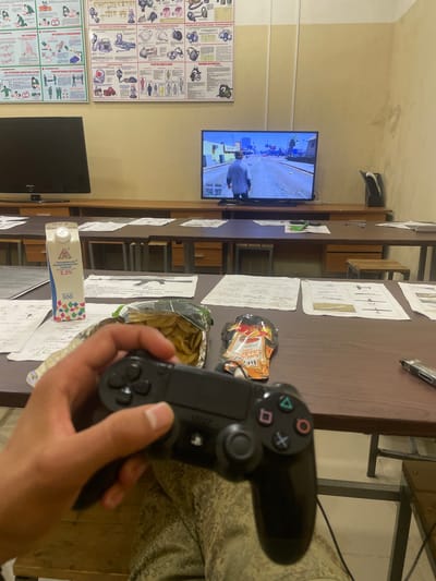 The image depicts a scene indoors, likely in a classroom or communal area, with a person holding a video game controller and playing a game on a television screen. The television displays a scene from the video game Grand Theft Auto V, showing a character walking down a street in a city. On the table in front of the person are a carton of milk and bags of snacks, suggesting a casual leisure activity. The room appears to be furnished with desks and chairs, and educational posters are visible on the wall, some of which detail safety procedures or technical information. The overall atmosphere is relaxed and informal. The location is identified as Оредеж, Russia.