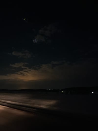 The image depicts a night scene in Gol'yany, Russia. The left side of the image shows dark sky with scattered clouds and a few distant lights, possibly from buildings or streetlights. The right side features a blurred and dark interior, likely a window view from inside a building or vehicle. The vertical black line suggests a window frame. The overall scene conveys an impression of limited visibility in the darkness.