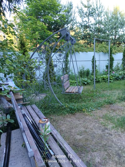 In the city of Космос-2 Снт, Russia, a garden scene unfolds under what appears to be daylight. A metal swing set with a wooden seat stands prominently in the center of the frame, its ornate scrollwork suggesting a decorative design. To the left, a wooden bench with various metal objects and a can of paint rests upon it, hinting at ongoing projects or stored materials. The background is dominated by lush greenery, including trees and a white fence that delineates the property. The ground is covered in grass and patches of dry earth, with scattered wildflowers adding touches of color. The overall impression is one of a lived-in, slightly untidy outdoor space.