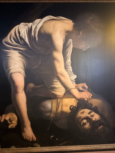 The image depicts a painting of a young man in a white robe standing over a decapitated giant. The young man's left leg is bent and placed on the giant's chest, and he is holding the giant's severed head by its hair with both hands. The giant's eyes are wide open, and his mouth is agape in a grimace. A drop of blood is visible on his forehead. The painting is lit dramatically, with the figures emerging from a dark background. The scene is interpreted as David and Goliath. The painting is located in the Iglesia de San Andrés in Mazarrón, Spain.