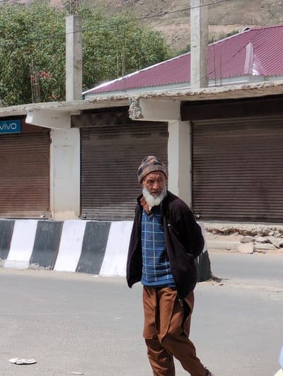 An elderly man with a white beard and a checkered beanie walks on a paved road in Padum, India. He is wearing a dark jacket over a blue checkered sweater and brown pants. In the background, there are closed storefronts with brown metal roll-up doors, a white concrete structure with exposed rebar, and a building with a reddish-brown metal roof. A striped barrier with black and white stripes is present in the foreground, separating the road from the sidewalk area where the storefronts are located. Green trees are visible behind the structures, and a rocky hillside forms the backdrop. The lighting suggests it is daytime, likely sunny, with shadows present.