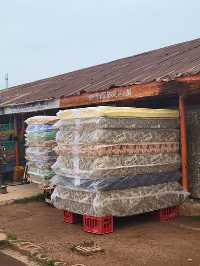 The image depicts an outdoor scene in the city of Tangu, Nigeria, likely in front of a business establishment. The foreground is dominated by stacks of mattresses, many of which are wrapped in clear plastic packaging. These mattresses are placed on top of red Coca-Cola crates, which elevate them from the dirt and gravel ground. Some of the mattresses have visible labels, such as "Ultrafoam."

Behind the mattresses, a building with a weathered, corrugated metal roof is visible. The building appears to be a shop or distributor, as evidenced by signage. One sign reads "EJIRE COLLECTION (NIG.) Dealer and Supplies of all types of Foreign Building Materials". Another sign, partially obscured, reads "CHRIS INTEGRATED RESOURCES DISTRIBUTOR". The building's exterior walls are a pale color, and there is an orange support beam visible.

To the left of the building, there are some items that suggest the shop's inventory or services. These include what appear to be some other building materials, possibly colored planks or panels, and a yellow jerrycan. Hanging from a string are some black circular objects, their purpose unclear. The sky in the background is overcast, with a faint suggestion of a telecommunications tower in the far distance. The overall environment appears to be a commercial area, likely a street-side market or shop front. There are no people visible in the image.