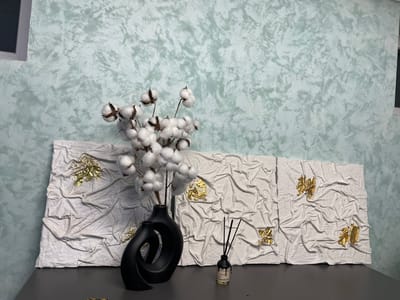The image depicts an indoor scene with a focus on decorative objects placed on a dark gray table. In the foreground, a striking black vase with a unique, stylized design holds several cotton stems with fluffy white cotton bolls. To the right of the vase sits a small, dark-labeled bottle with black reed diffuser sticks protruding from it.
Behind these objects, a triptych artwork is displayed. Each panel of the artwork features a textured white surface with gold leaf embellishments scattered across it. The texture of the white surface appears to mimic the look of crumpled fabric.
The background consists of a wall with a mottled, textured surface in shades of light blue and gray. A portion of a window frame is visible on the left side of the image.
The image was taken in Лесной Пейзаж Кп (шарапово), Russia.