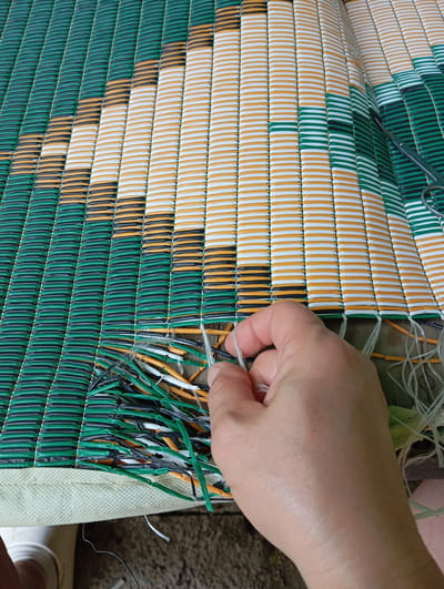 The image shows a close-up of a hand working on a woven mat. The mat is made of green, white, and orange plastic strips. The hand is holding a white thread, possibly for mending the mat. Some loose plastic strips and threads are visible on the edge of the mat. The image does not provide any information that can be used to determine the location in the city of Yukariyuz, Uzbekistan.