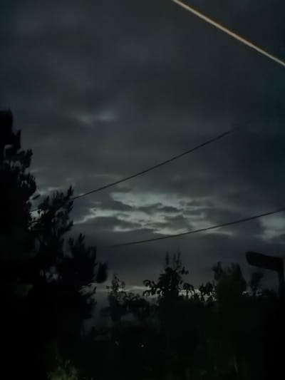 The image shows a nighttime scene, likely in a rural area, in the city of Novxanı, Azerbaijan. Dark, ominous clouds fill the sky. Two parallel wires stretch horizontally across the view. On the right side of the image, dark, indistinct shapes suggest trees and vegetation. A bright white line is present at the top left. The overall mood is somber and suggests an impending storm.